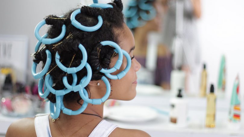 A imagem mostra uma mulher de costas com bigudinhos azuis no cabelo, um acessório usado para criar cachos definidos. Ela está em um ambiente que parece ser um salão de beleza ou uma penteadeira, com produtos capilares e espelhos ao fundo. O visual sugere um processo de finalização ou modelagem dos fios para obter cachos mais estruturados.
