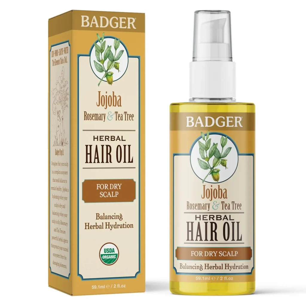 A imagem apresenta um frasco e uma caixa do Badger Herbal Hair Oil, um óleo capilar à base de jojoba, alecrim e tea tree. O produto é indicado para couro cabeludo seco, oferecendo hidratação equilibrada com ingredientes herbais. A embalagem destaca que é orgânico certificado pela USDA e tem um design clássico com tons dourados e ilustração botânica.